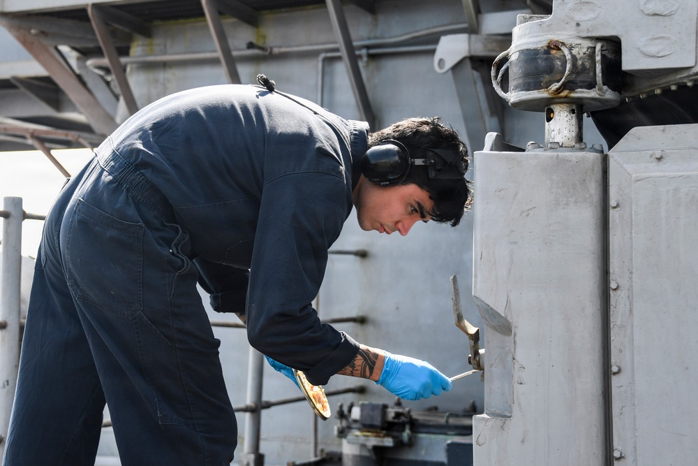 A young male technician or sailor in dark navy coveralls is performing maintenance work on heavy metal equipment, likely aboard a naval vessel. He is bent over and focused on manipulating components with tools while wearing hearing protection and blue nitrile gloves. The industrial metal surroundings and structural elements suggest a shipboard or heavy machinery environment.