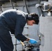 A young male technician or sailor in dark navy coveralls is performing maintenance work on heavy metal equipment, likely aboard a naval vessel. He is bent over and focused on manipulating components with tools while wearing hearing protection and blue nitrile gloves. The industrial metal surroundings and structural elements suggest a shipboard or heavy machinery environment.