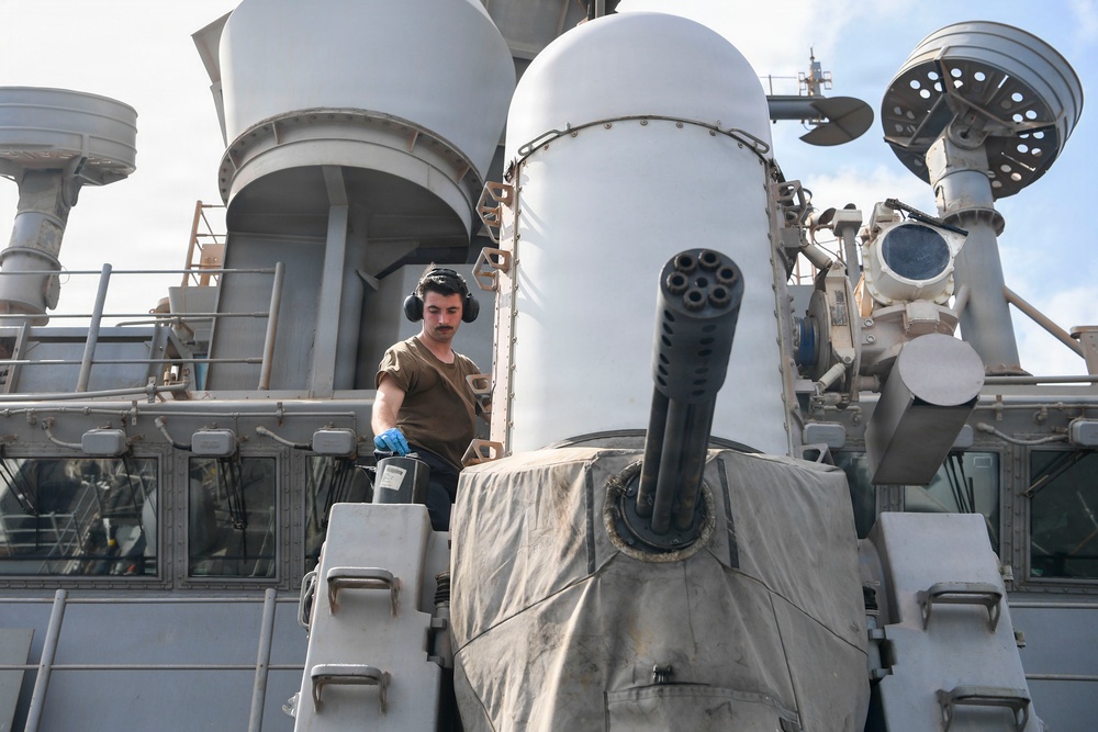 A military sailor or technician is working on the deck of a naval warship, positioned near a close-in weapon system (CIWS), likely a Phalanx system, identifiable by its distinctive multi-barrel rotary cannon and cylindrical radar dome. The personnel is wearing hearing protection and blue gloves, suggesting active maintenance or operational work. The ship's superstructure, including radar arrays and various sensor equipment, is visible in the background.