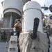 A military sailor or technician is working on the deck of a naval warship, positioned near a close-in weapon system (CIWS), likely a Phalanx system, identifiable by its distinctive multi-barrel rotary cannon and cylindrical radar dome. The personnel is wearing hearing protection and blue gloves, suggesting active maintenance or operational work. The ship's superstructure, including radar arrays and various sensor equipment, is visible in the background.