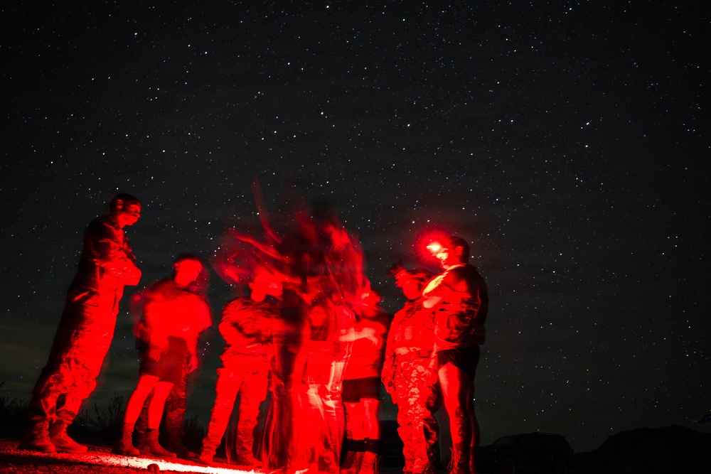 A group of individuals illuminated by red light stand outdoors at night under a star-filled sky. The long-exposure photograph creates motion blur among the figures, giving them a ghostly appearance. The subjects appear to be wearing military-style uniforms or tactical gear.