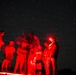 A group of individuals illuminated by red light stand outdoors at night under a star-filled sky. The long-exposure photograph creates motion blur among the figures, giving them a ghostly appearance. The subjects appear to be wearing military-style uniforms or tactical gear.