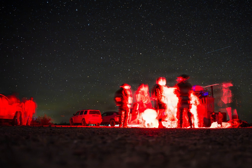 A long-exposure night photograph showing a group of people gathered around a bright red light source outdoors under a clear, star-filled sky. The long exposure has caused motion blur on the figures, giving them a ghostly, ethereal appearance. At least one vehicle is visible in the background, suggesting a remote outdoor location.