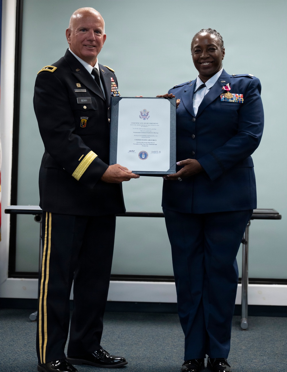 Two military officers are posing together while jointly holding a framed Certificate of Retirement from the United States Air Force. The officer on the left wears a U.S. Army dress uniform with the name tag 'BERRY,' while the officer on the right wears a U.S. Air Force dress uniform adorned with multiple medals and ribbons. The ceremony appears to be an official military retirement presentation.