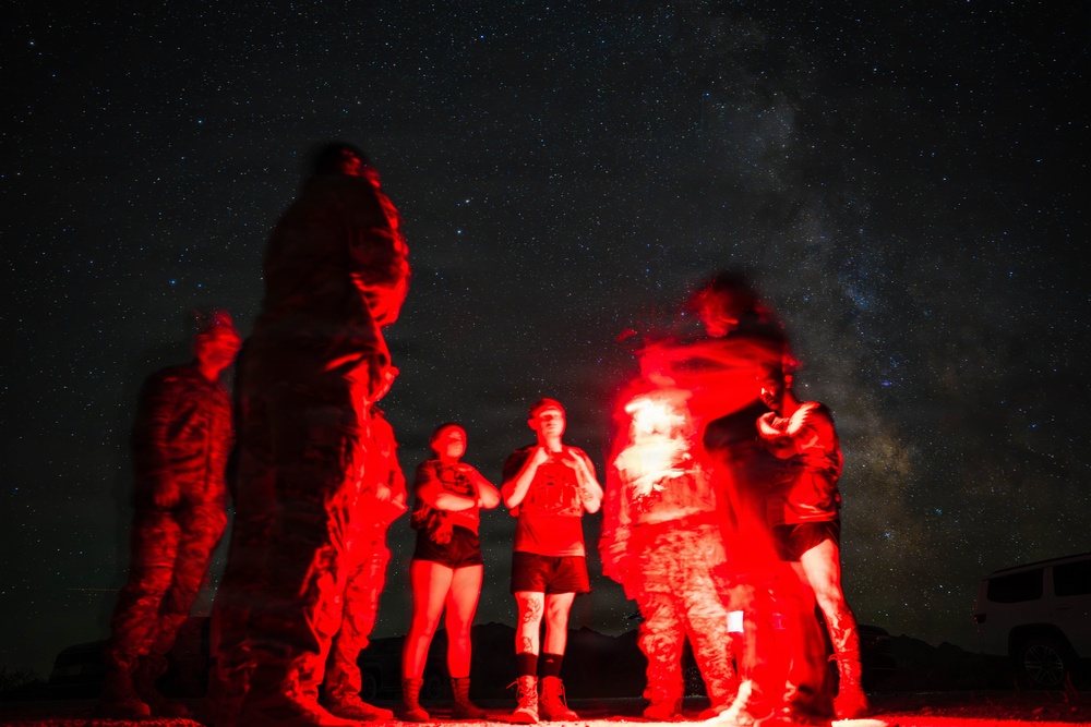 A group of people are gathered outdoors at night, illuminated by a red flare or signal light held by one of the individuals. Several people appear to be wearing military-style camouflage uniforms while others are in civilian clothing. The scene is set against a dramatic starry night sky with the Milky Way visible.