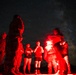 A group of people are gathered outdoors at night, illuminated by a red flare or signal light held by one of the individuals. Several people appear to be wearing military-style camouflage uniforms while others are in civilian clothing. The scene is set against a dramatic starry night sky with the Milky Way visible.