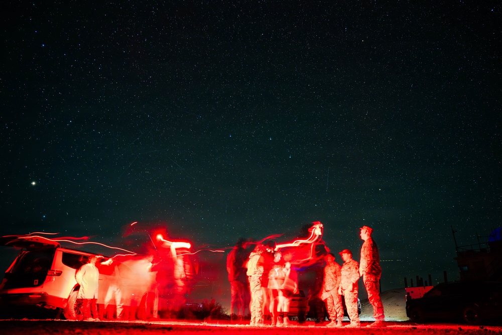 A long-exposure night photograph showing a group of individuals illuminated by red light against a star-filled sky. The red lighting and motion blur from the extended exposure create ghostly, streaked silhouettes of people gathered around vehicles. The setting appears to be an open, remote outdoor area at night.