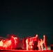 A long-exposure night photograph showing a group of individuals illuminated by red light against a star-filled sky. The red lighting and motion blur from the extended exposure create ghostly, streaked silhouettes of people gathered around vehicles. The setting appears to be an open, remote outdoor area at night.