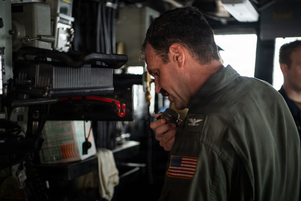 A U.S. military officer in a green flight suit is speaking into a handheld radio or microphone aboard what appears to be a naval vessel's bridge or command area. He has a U.S. flag patch on his right shoulder and what appears to be aviator wings on his chest, indicating a naval aviator rank. A second person is partially visible in the background near a window.