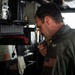 A U.S. military officer in a green flight suit is speaking into a handheld radio or microphone aboard what appears to be a naval vessel's bridge or command area. He has a U.S. flag patch on his right shoulder and what appears to be aviator wings on his chest, indicating a naval aviator rank. A second person is partially visible in the background near a window.
