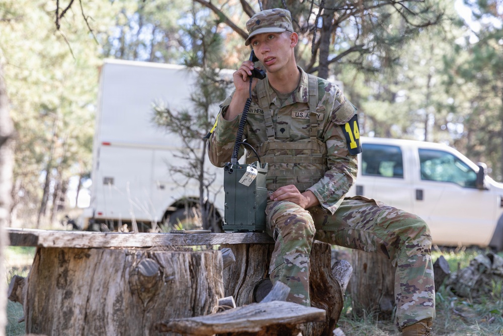 A U.S. Army soldier in MultiCam (OCP) camouflage uniform sits on a tree stump in a wooded outdoor area, using a military field radio handset connected to a portable radio unit. The soldier wears a tactical vest and displays the number '14' on a yellow patch, suggesting a training exercise or competition event. White military or government vehicles are visible in the background among pine trees.