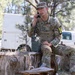 A U.S. Army soldier in MultiCam (OCP) camouflage uniform sits on a tree stump in a wooded outdoor area, using a military field radio handset connected to a portable radio unit. The soldier wears a tactical vest and displays the number '14' on a yellow patch, suggesting a training exercise or competition event. White military or government vehicles are visible in the background among pine trees.