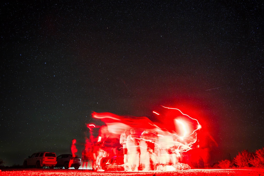 A long-exposure night photography shot showing multiple people illuminated by red light against a star-filled sky. The extended shutter speed has created motion blur and light trails, giving the figures a ghostly, ethereal appearance. Parked vehicles are visible on the left side of the frame.
