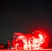 A long-exposure night photography shot showing multiple people illuminated by red light against a star-filled sky. The extended shutter speed has created motion blur and light trails, giving the figures a ghostly, ethereal appearance. Parked vehicles are visible on the left side of the frame.