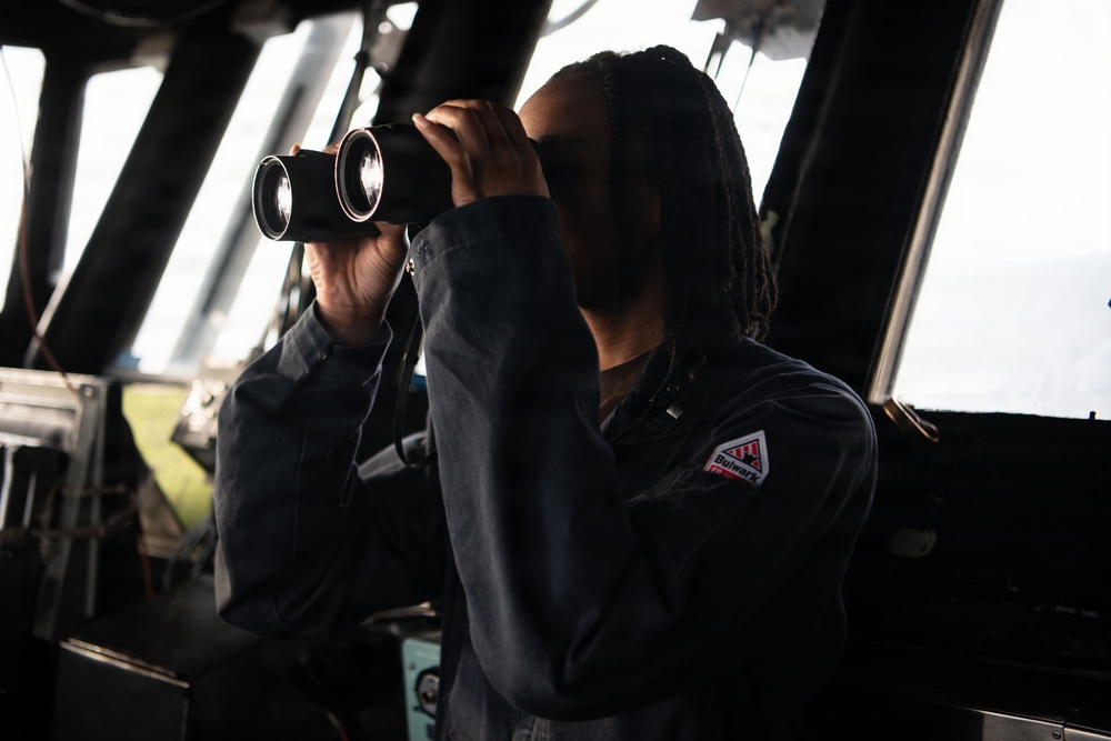 A person wearing a dark navy coverall uniform is using large binoculars while standing on what appears to be the bridge of a ship. A second individual in similar attire is visible in the background. The setting is clearly a maritime vessel's bridge or wheelhouse, with navigation equipment and large windows visible.