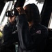 A person wearing a dark navy coverall uniform is using large binoculars while standing on what appears to be the bridge of a ship. A second individual in similar attire is visible in the background. The setting is clearly a maritime vessel's bridge or wheelhouse, with navigation equipment and large windows visible.