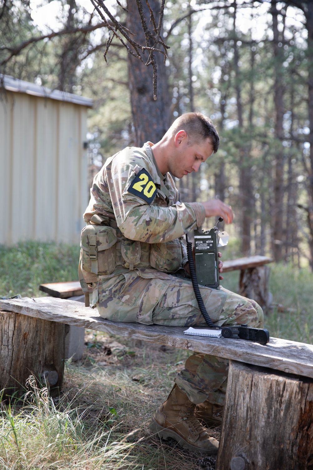 A soldier in OCP (Operational Camouflage Pattern) uniform, marked with the number '20' on a black and yellow patch, sits on a wooden bench in a forested outdoor area. He is operating or configuring what appears to be a military field radio (likely an AN/PRC-type radio), with a handset cord visible. A spiral notebook and what appears to be a pen or stylus are placed on the bench beside him.