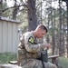 A soldier in OCP (Operational Camouflage Pattern) uniform, marked with the number '20' on a black and yellow patch, sits on a wooden bench in a forested outdoor area. He is operating or configuring what appears to be a military field radio (likely an AN/PRC-type radio), with a handset cord visible. A spiral notebook and what appears to be a pen or stylus are placed on the bench beside him.
