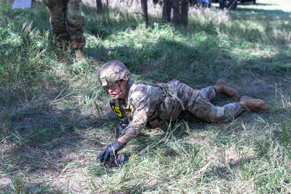A soldier in full camouflage uniform and helmet is performing a low crawl exercise on grassy terrain, wearing a yellow competition number bib (number 5). The activity appears to be part of a military training exercise or competition event, with another uniformed individual visible in the background. The soldier is prone on the ground, using their arms to propel forward through the grass.