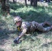 A soldier in full camouflage uniform and helmet is performing a low crawl exercise on grassy terrain, wearing a yellow competition number bib (number 5). The activity appears to be part of a military training exercise or competition event, with another uniformed individual visible in the background. The soldier is prone on the ground, using their arms to propel forward through the grass.