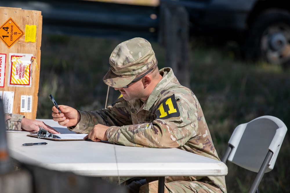 A U.S. Army soldier in Operational Camouflage Pattern (OCP) uniform is seated at a folding table, writing on documents attached to a clipboard. The soldier wears a patch with the number '11' on the left sleeve, consistent with 11th Corps or a related unit designation. In the background, a wooden crate bears an explosive hazard placard rated '1.4 S', indicating a low-hazard explosive storage or transport context.
