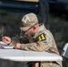 A U.S. Army soldier in Operational Camouflage Pattern (OCP) uniform is seated at a folding table, writing on documents attached to a clipboard. The soldier wears a patch with the number '11' on the left sleeve, consistent with 11th Corps or a related unit designation. In the background, a wooden crate bears an explosive hazard placard rated '1.4 S', indicating a low-hazard explosive storage or transport context.