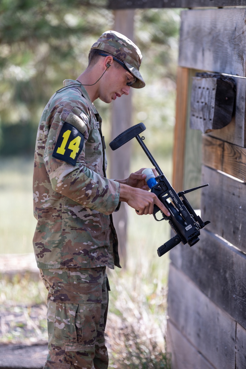 A soldier in OCP (Operational Camouflage Pattern) uniform wearing number '14' on their left arm is handling what appears to be a grenade launcher or specialized weapon system near a wooden wall structure. The individual is wearing protective eyewear pushed up on their face and has an earpiece in. This appears to be a military training exercise or competition event.