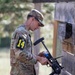 A soldier in OCP (Operational Camouflage Pattern) uniform wearing number '14' on their left arm is handling what appears to be a grenade launcher or specialized weapon system near a wooden wall structure. The individual is wearing protective eyewear pushed up on their face and has an earpiece in. This appears to be a military training exercise or competition event.