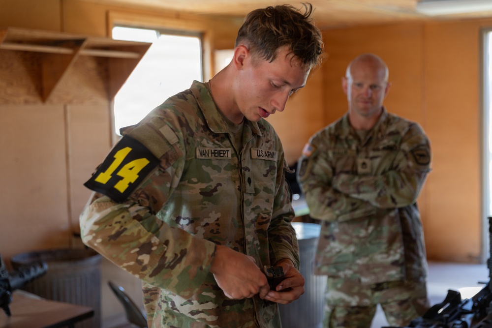 Two U.S. Army soldiers are inside a building, with the foreground soldier (name tape reading 'VAN HEBERT') wearing a yellow '14' competition number patch on his left arm and handling a small object. A second soldier stands in the background with arms crossed, appearing to observe. The setting and competition number suggest this may be a military skills competition or evaluation event.