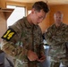 Two U.S. Army soldiers are inside a building, with the foreground soldier (name tape reading 'VAN HEBERT') wearing a yellow '14' competition number patch on his left arm and handling a small object. A second soldier stands in the background with arms crossed, appearing to observe. The setting and competition number suggest this may be a military skills competition or evaluation event.