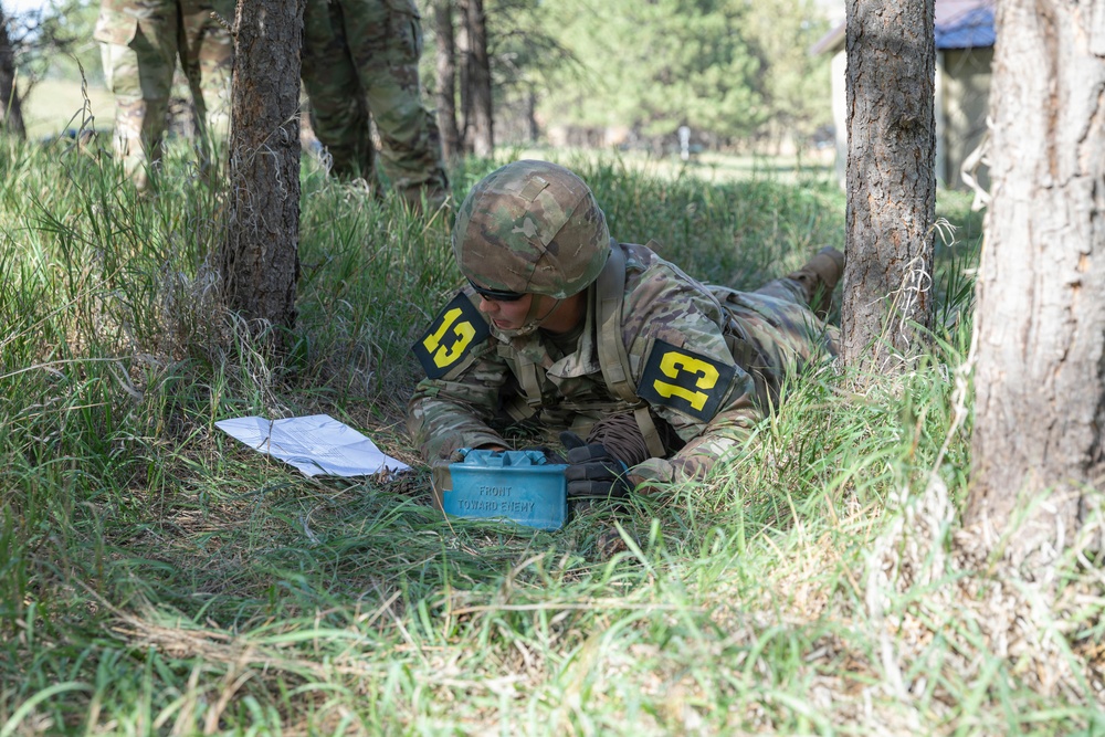 A soldier wearing camouflage uniform and helmet, marked with the number '13' on both shoulders, is lying prone on the ground in a wooded training area. The soldier is examining or working with a blue training device labeled 'FRONT TOWARD ENEMY,' which is a replica of an M18A1 Claymore mine used for training purposes. Papers are visible nearby, suggesting this is a graded or evaluated military training exercise.