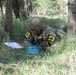 A soldier wearing camouflage uniform and helmet, marked with the number '13' on both shoulders, is lying prone on the ground in a wooded training area. The soldier is examining or working with a blue training device labeled 'FRONT TOWARD ENEMY,' which is a replica of an M18A1 Claymore mine used for training purposes. Papers are visible nearby, suggesting this is a graded or evaluated military training exercise.