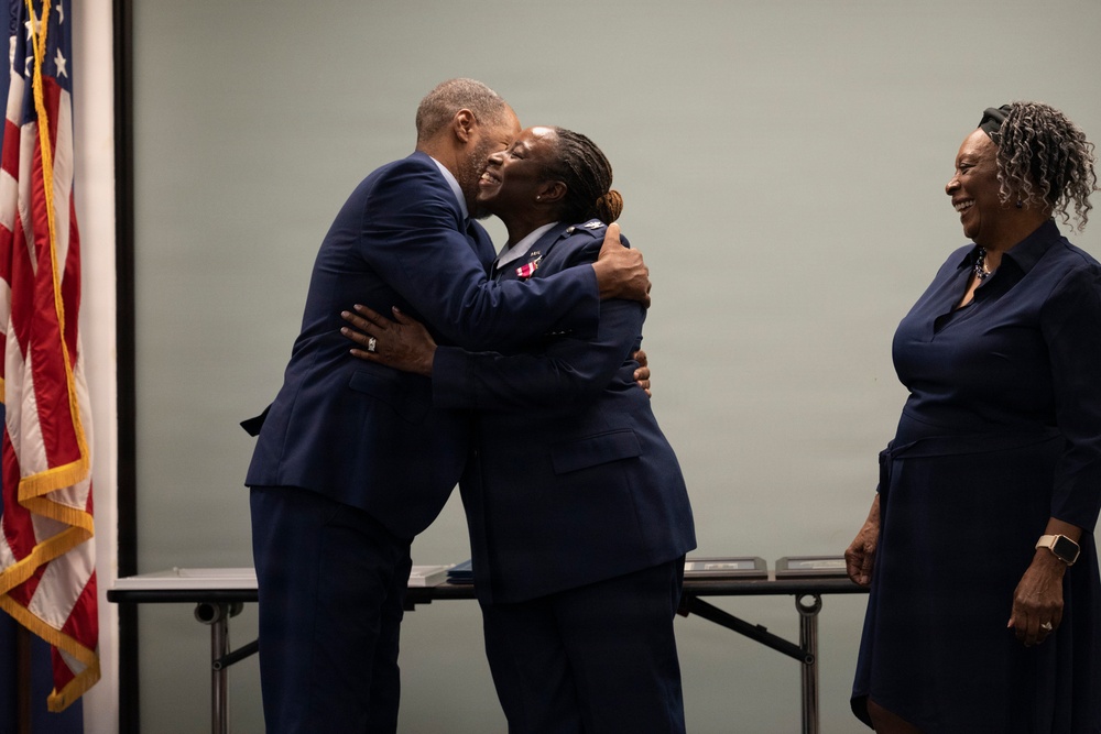 A military ceremony appears to be taking place, with a person in a U.S. Air Force uniform receiving a congratulatory embrace from a man in a navy suit. A third person in a navy dress stands nearby smiling. An American flag with gold fringe is visible in the background, and what appear to be award plaques or folders are on a table behind them.