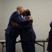 A military ceremony appears to be taking place, with a person in a U.S. Air Force uniform receiving a congratulatory embrace from a man in a navy suit. A third person in a navy dress stands nearby smiling. An American flag with gold fringe is visible in the background, and what appear to be award plaques or folders are on a table behind them.
