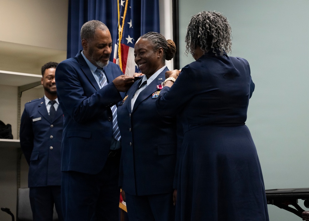 A military promotion ceremony is taking place, with a uniformed U.S. Air Force officer being pinned on both shoulders simultaneously by two civilians, likely family members. The officer is smiling broadly, indicating a joyful milestone moment. An American flag and a young airman in uniform are visible in the background.
