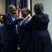 A military promotion ceremony is taking place, with a uniformed U.S. Air Force officer being pinned on both shoulders simultaneously by two civilians, likely family members. The officer is smiling broadly, indicating a joyful milestone moment. An American flag and a young airman in uniform are visible in the background.