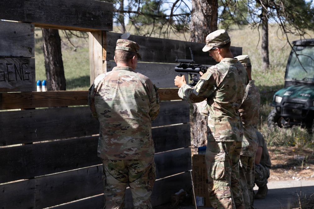 Two military personnel in OCP (Operational Camouflage Pattern) uniforms are positioned at a wooden pallet barrier during what appears to be a training exercise. One soldier is aiming a rifle equipped with an optic sight through a gap in the barrier, while the other observes. The setting is an outdoor training range with trees and a utility vehicle visible in the background.