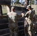 Two military personnel in OCP (Operational Camouflage Pattern) uniforms are positioned at a wooden pallet barrier during what appears to be a training exercise. One soldier is aiming a rifle equipped with an optic sight through a gap in the barrier, while the other observes. The setting is an outdoor training range with trees and a utility vehicle visible in the background.