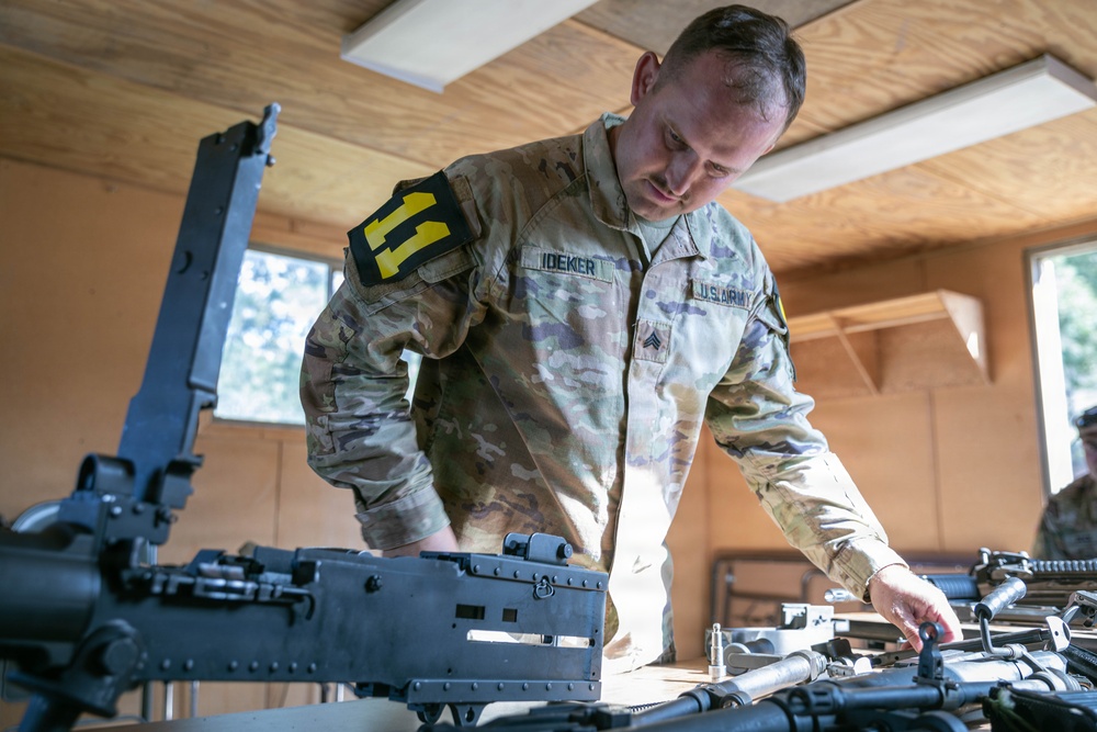 A U.S. Army soldier, identified by name tape as 'IDEKER' with the rank of Sergeant, is working with military weapons on a table inside a wooden structure. The soldier is wearing Operational Camouflage Pattern (OCP) uniform with a unit patch on the left shoulder featuring the number '11' on a black and yellow background. The activity appears to be weapons maintenance, inspection, or inventory.