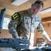 A U.S. Army soldier, identified by name tape as 'IDEKER' with the rank of Sergeant, is working with military weapons on a table inside a wooden structure. The soldier is wearing Operational Camouflage Pattern (OCP) uniform with a unit patch on the left shoulder featuring the number '11' on a black and yellow background. The activity appears to be weapons maintenance, inspection, or inventory.