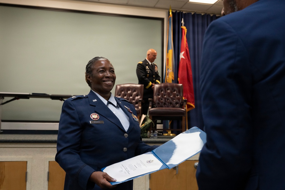 A U.S. Air Force officer in dress blues is smiling while receiving or exchanging an official document or certificate during what appears to be a military ceremony. In the background, a U.S. Army officer in dress greens stands near a stage with military flags, including one bearing a single star. The setting appears to be an indoor military ceremony or award presentation.