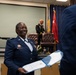 A U.S. Air Force officer in dress blues is smiling while receiving or exchanging an official document or certificate during what appears to be a military ceremony. In the background, a U.S. Army officer in dress greens stands near a stage with military flags, including one bearing a single star. The setting appears to be an indoor military ceremony or award presentation.
