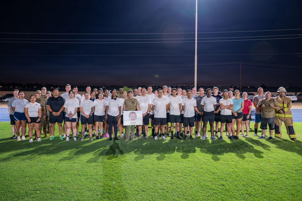 A large group of approximately 40 individuals poses for a group photo on a lit athletic track field at night. The group appears to be a mix of military personnel, civilians, and at least one firefighter in full turnout gear. A sign or placard is displayed on a stand in the center of the group, suggesting an organized commemorative or unit event.