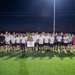 A large group of approximately 40 individuals poses for a group photo on a lit athletic track field at night. The group appears to be a mix of military personnel, civilians, and at least one firefighter in full turnout gear. A sign or placard is displayed on a stand in the center of the group, suggesting an organized commemorative or unit event.