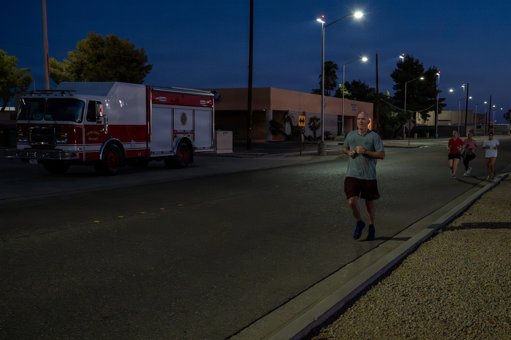 A group of people are jogging along a road during early morning or evening hours under artificial street lighting. A red and white United States Air Force fire truck is parked on the left side of the road. The setting appears to be a military base, suggested by the fire truck markings and institutional building architecture.