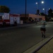 A group of people are jogging along a road during early morning or evening hours under artificial street lighting. A red and white United States Air Force fire truck is parked on the left side of the road. The setting appears to be a military base, suggested by the fire truck markings and institutional building architecture.