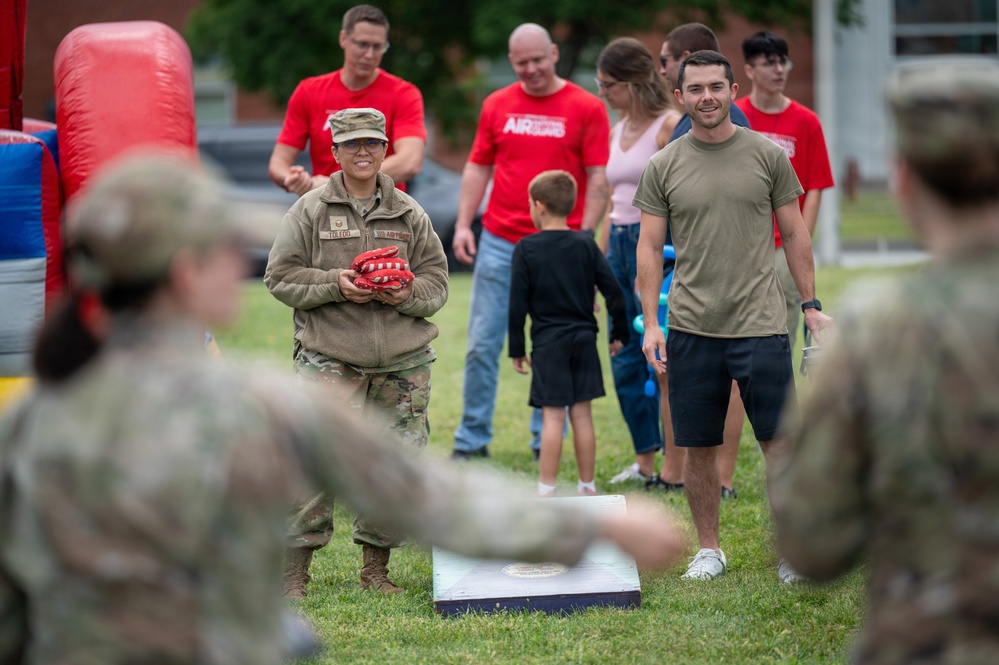 A military outdoor gathering or family day event where service members and civilians are participating in recreational activities on a grass field. A soldier in camouflage uniform holding red bean bags is preparing to play cornhole, with a decorated cornhole board visible in the foreground. A mix of uniformed personnel and civilians in red shirts, including children, are present in the background.