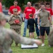 A military outdoor gathering or family day event where service members and civilians are participating in recreational activities on a grass field. A soldier in camouflage uniform holding red bean bags is preparing to play cornhole, with a decorated cornhole board visible in the foreground. A mix of uniformed personnel and civilians in red shirts, including children, are present in the background.
