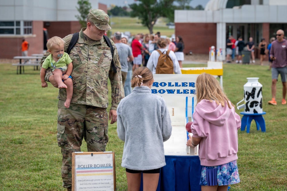 A military service member in camouflage uniform holds a young infant while standing near a 'Roller Bowler' carnival game at an outdoor family event. Two girls with their backs to the camera appear to be playing or observing the game. The setting appears to be a military base family day or community event on a grassy field.