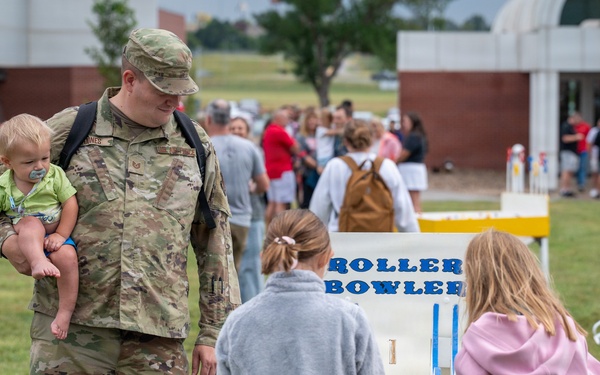 A military service member in camouflage uniform holds a young infant while standing near a 'Roller Bowler' carnival game at an outdoor family event. Two girls with their backs to the camera appear to be playing or observing the game. The setting appears to be a military base family day or community event on a grassy field.